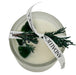 White candle cup with green stones embedded and ribbon with juniper tied on top.