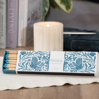 Box of decorative matches with a floral design on a table with a lit candle in the background.