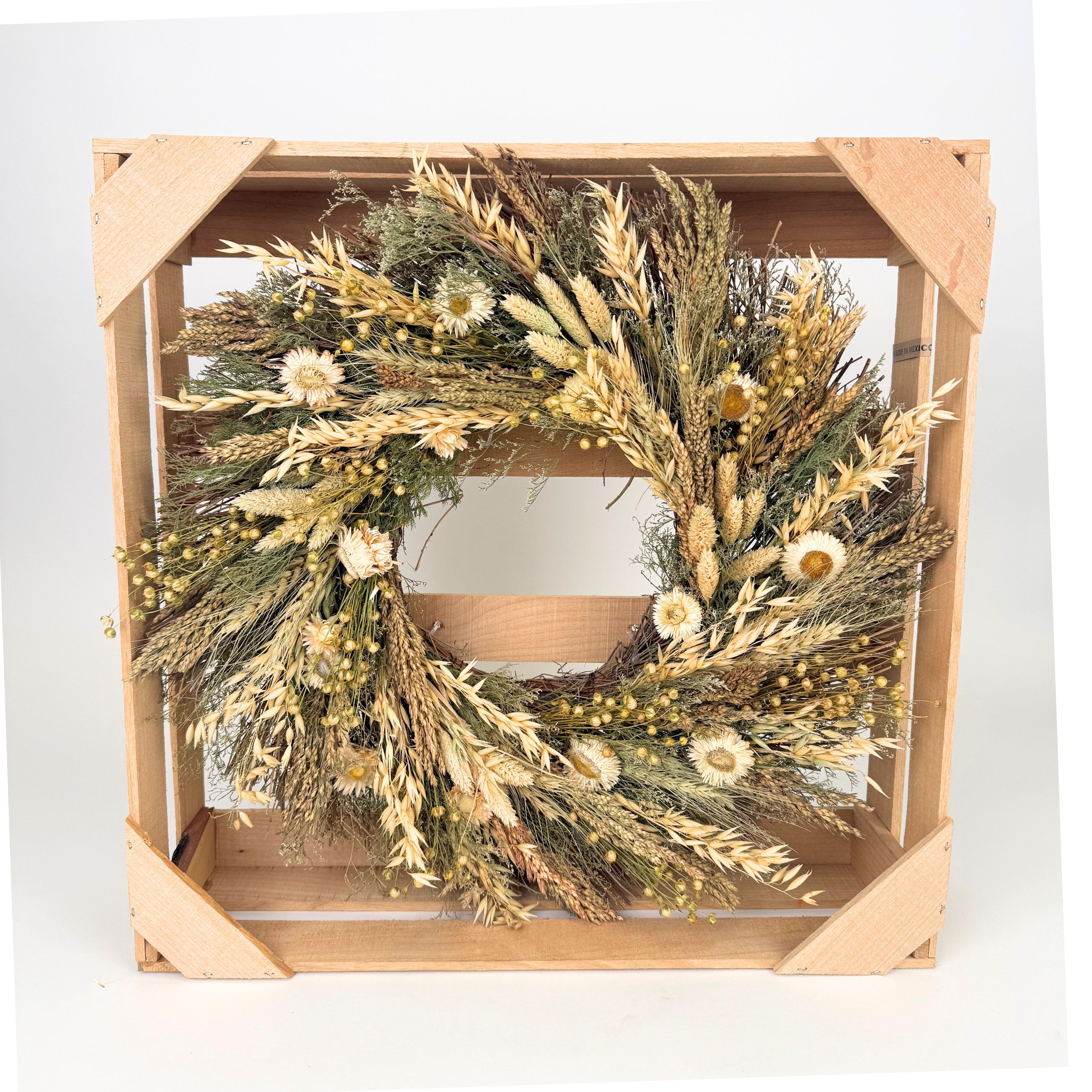 A dried flower wreath made of natural beige, brown, and green botanicals, displayed inside a square light wood crate on a white background.