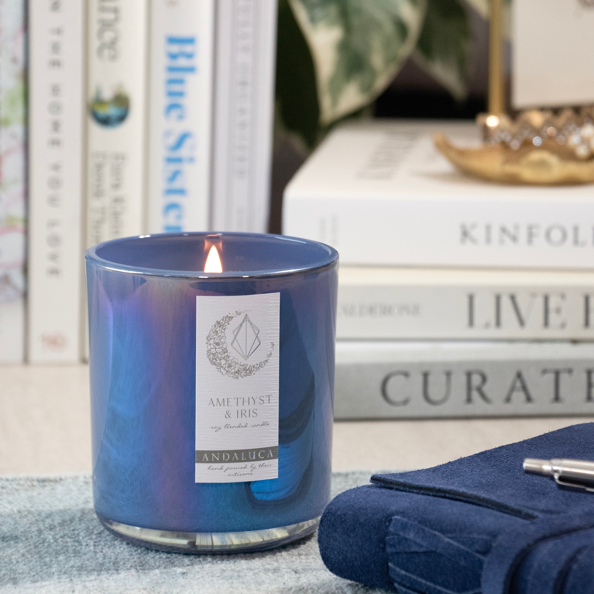 A lit blue glass candle jar with a white label reading 'AMETHYST & IRIS' and 'ANDALUCIA', styled on a tabletop with books, a folded navy cloth, and a pen in the background.