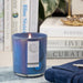 A lit blue glass candle jar with a white label reading 'AMETHYST & IRIS' and 'ANDALUCIA', styled on a tabletop with books, a folded navy cloth, and a pen in the background.