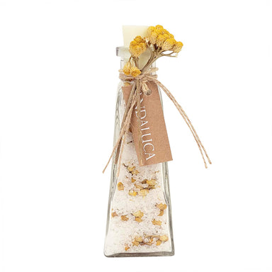 A clear glass bottle filled with white bath salts and dried yellow flowers, tied with twine and a kraft paper tag that reads 'BATH SALTS DAUJA'.