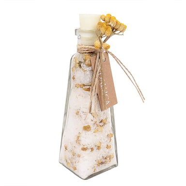 A clear glass bottle with a pointed bottom, filled with white bath salts or sugar crystals mixed with dried yellow flowers. The bottle is sealed with a white stopper, tied with twine, and has a kraft paper tag attached, all on a plain white background.