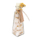 A clear glass bottle with a pointed bottom, filled with white bath salts or sugar crystals mixed with dried yellow flowers. The bottle is sealed with a white stopper, tied with twine, and has a kraft paper tag attached, all on a plain white background.
