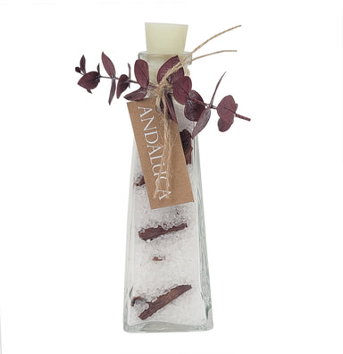 A clear glass decorative bottle with a cream-colored stopper, filled with white salt crystals and small brown bark pieces, tied with twine and a kraft tag reading ANDALUCIA, and adorned with dark burgundy eucalyptus leaves on a white background.