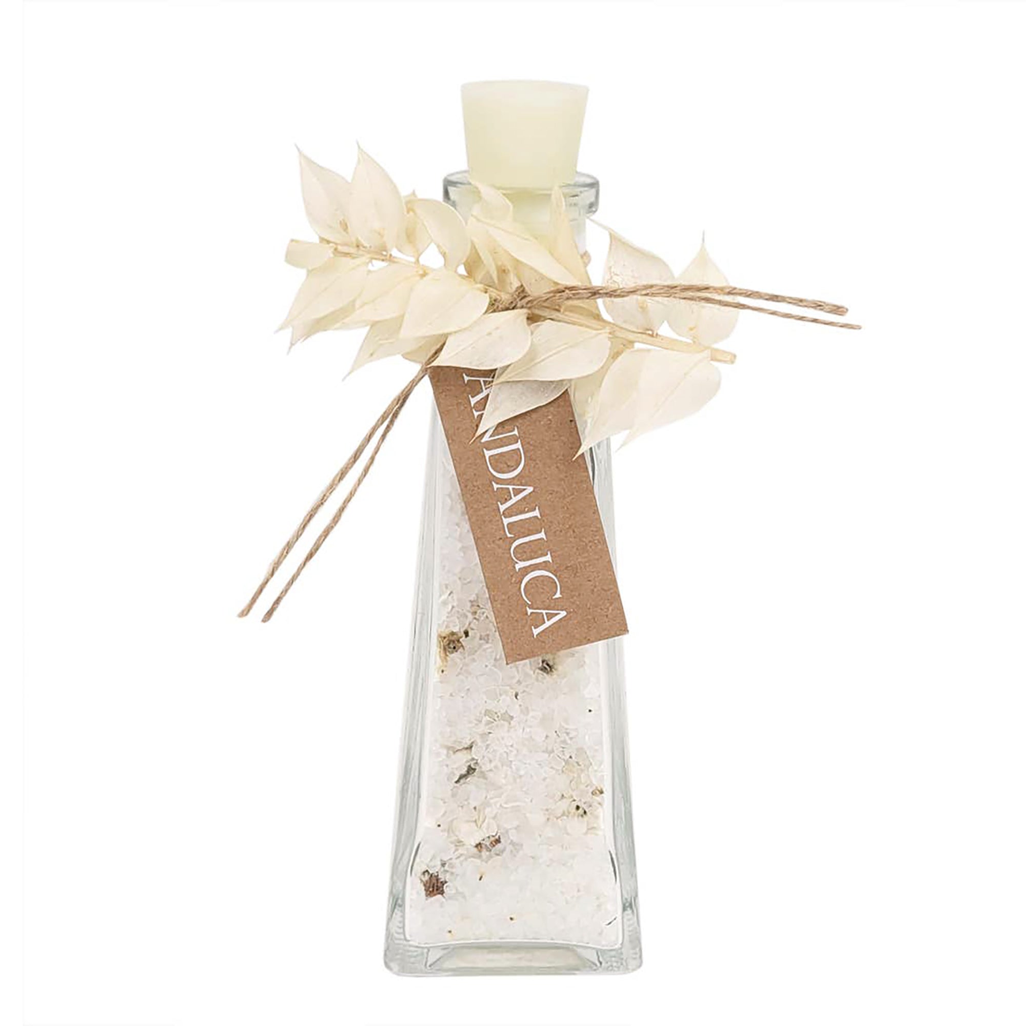 A clear glass bottle filled with white salt crystals and small dried botanical pieces, topped with a cream-colored wax seal, decorated with dried white leaves tied with twine and a brown paper tag reading 'ITALICA'.