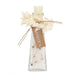 A clear glass bottle filled with white salt crystals and small dried botanical pieces, topped with a cream-colored wax seal, decorated with dried white leaves tied with twine and a brown paper tag reading 'ITALICA'.