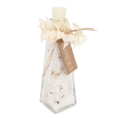 A clear glass bottle with a white wax seal on top, filled with white bath salts or sand, decorated with dried beige leaves, twine, and a brown paper tag that reads 'ANDALUCIA'.