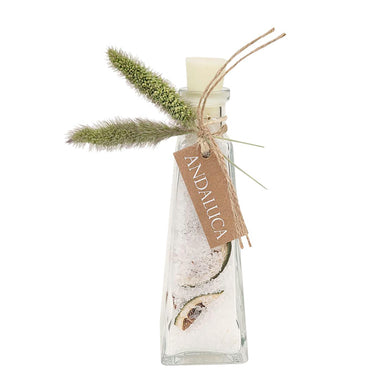 A clear glass bottle filled with white salt crystals and dried green botanical pieces, sealed with a white plug and tied with twine. A kraft paper tag reads 'ANDALUCIA' and is attached with green grass and dried stems against a plain white background.