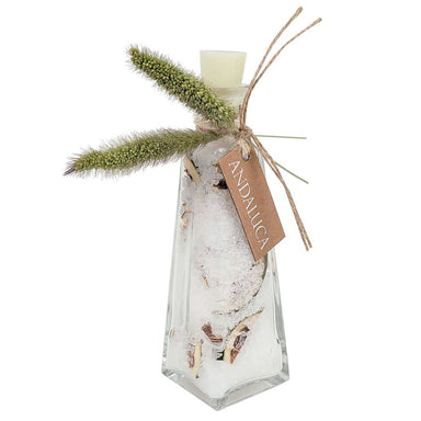 A clear glass bottle with a geometric shape, filled with white salt crystals and dried botanical pieces, topped with a white wax melt. It is decorated with two green dried grass stalks and a brown paper tag that reads 'ANDALUCA'.