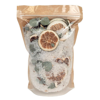 A clear resealable stand-up pouch with a kraft paper top, filled with coarse white crystals and dried botanicals including green leaves and dried citrus slices, photographed on a plain white background.