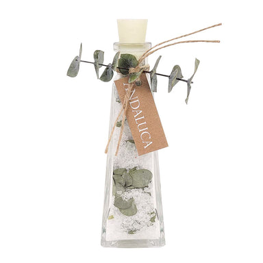 A clear glass bottle with a cream-colored stopper, filled with white sand or salt, green eucalyptus leaves, and a brown kraft paper tag tied with twine. The tag reads 'ANDALUCIA'.
