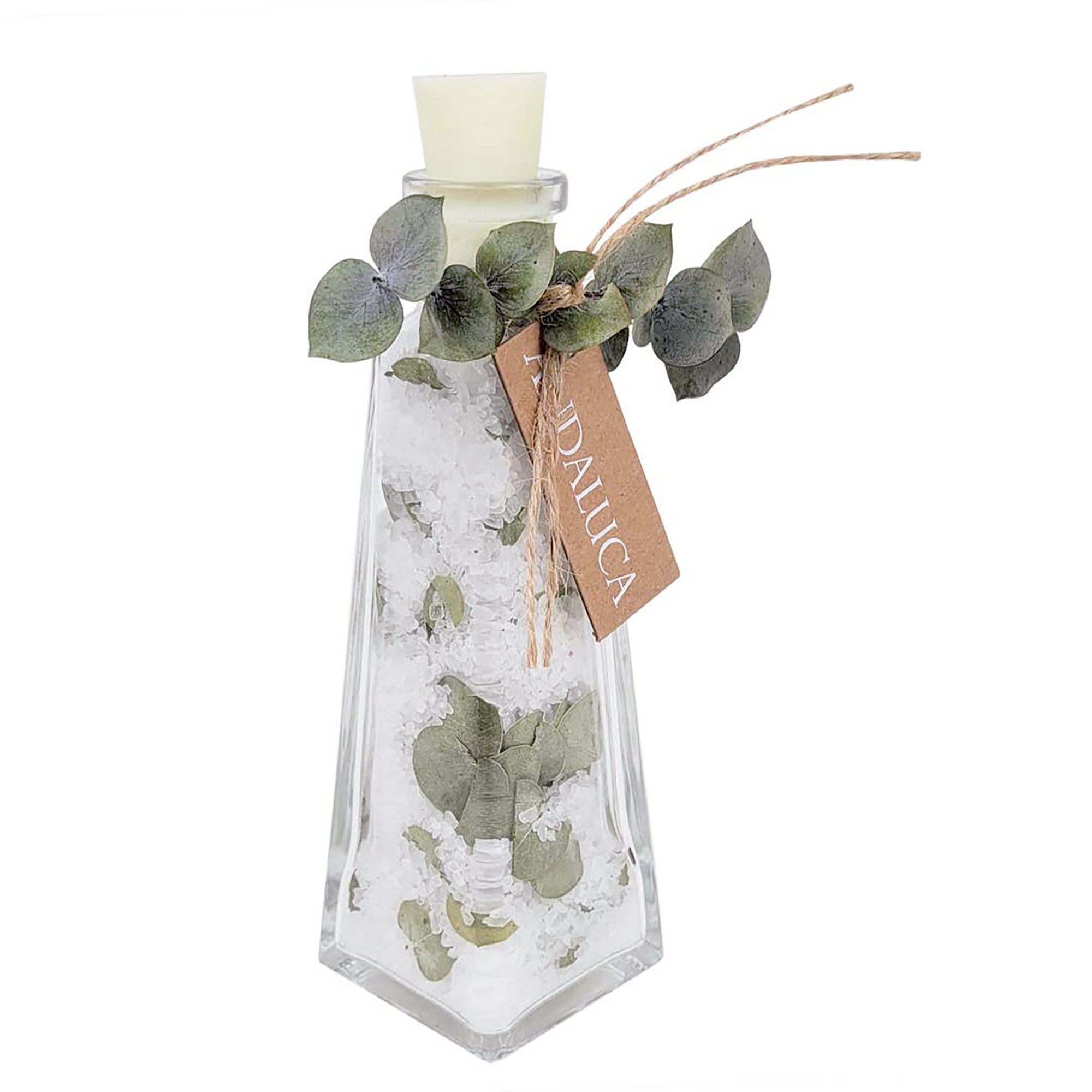 A clear glass decorative bottle with a white stopper, filled with white crystals or salt, decorated with green eucalyptus leaves and a brown paper tag that reads 'EUDALICA'.