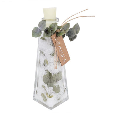 A clear glass decorative bottle with a white stopper, filled with white crystals or salt, decorated with green eucalyptus leaves and a brown paper tag that reads 'EUDALICA'.
