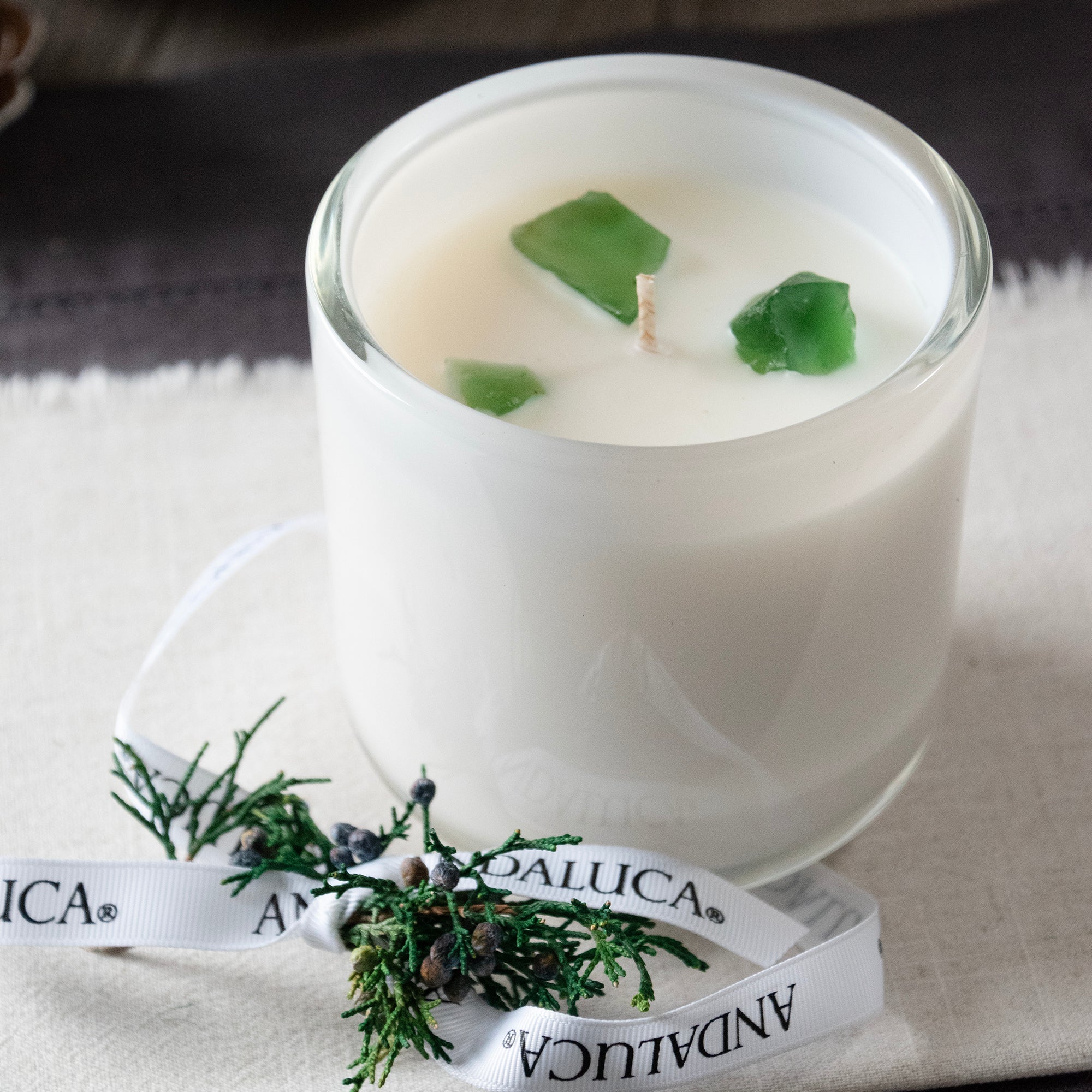 Unlite white glass candle vessel with juniper berry sprig tied into Andaluca branded white grossgrain ribbon, placed on the side reveling 3 green translucent stones surounding the wick. Stones do not need to be removed while burning.