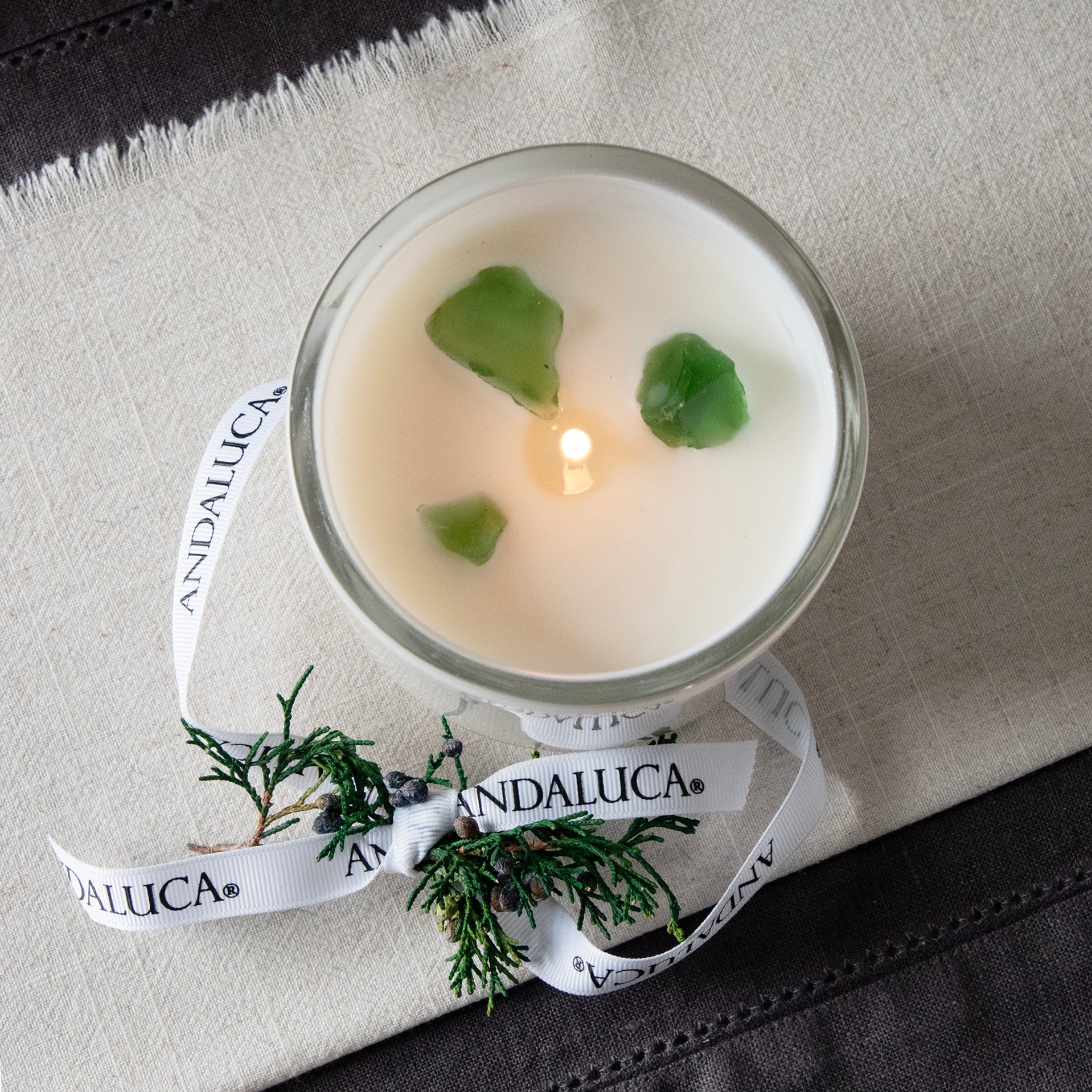 Over head view of lite white glass candle vessel with juniper berry sprig tied into Andaluca branded white grossgrain ribbon, placed on the side reveling 3 green translucent stones surounding the wick. Stones do not need to be removed while burning.