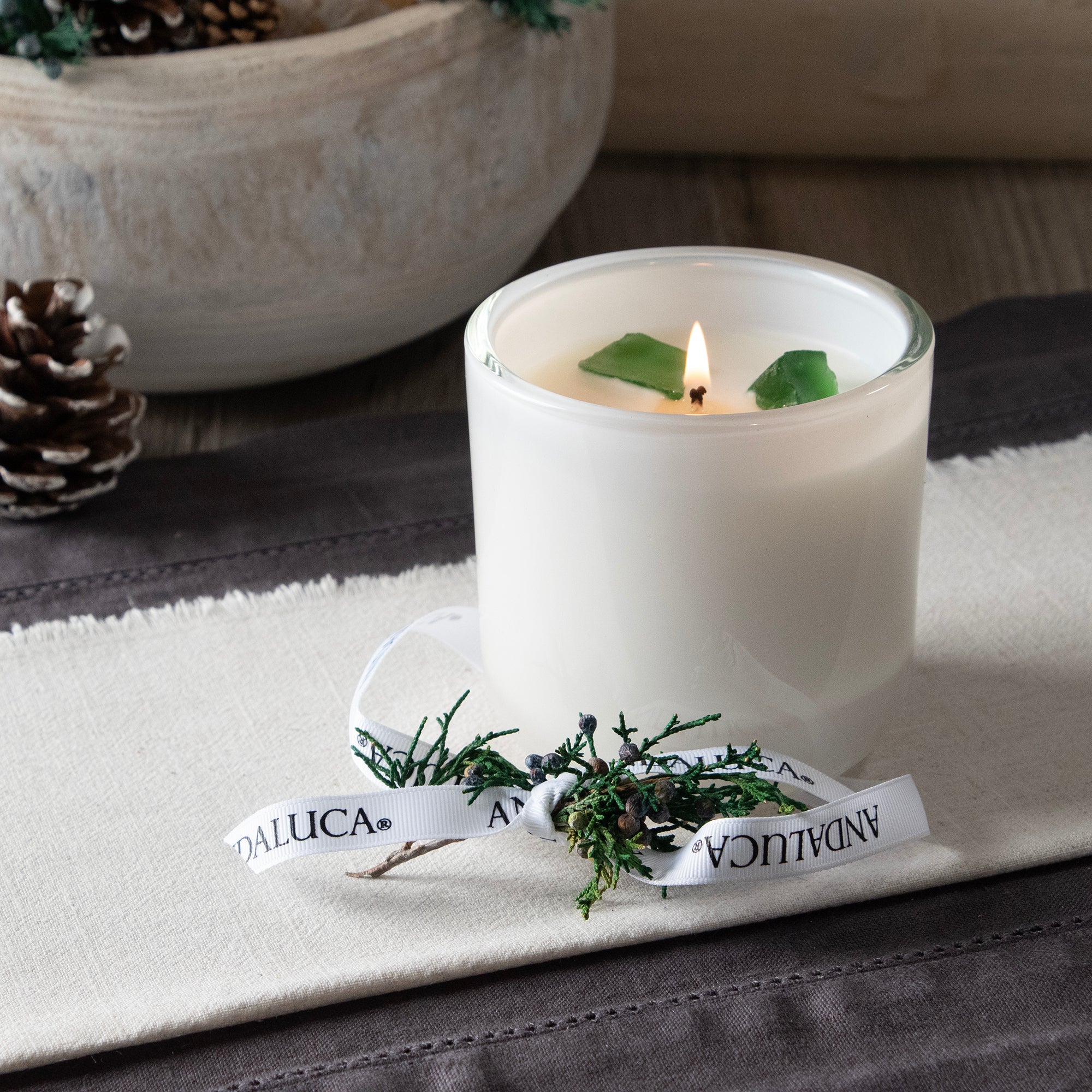 Lite white glass candle vessel with juniper berry sprig tied into Andaluca branded white grossgrain ribbon, placed on the side reveling 3 green translucent stones surounding the wick. Stones do not need to be removed while burning.