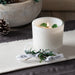 Lite white glass candle vessel with juniper berry sprig tied into Andaluca branded white grossgrain ribbon, placed on the side reveling 3 green translucent stones surounding the wick. Stones do not need to be removed while burning.