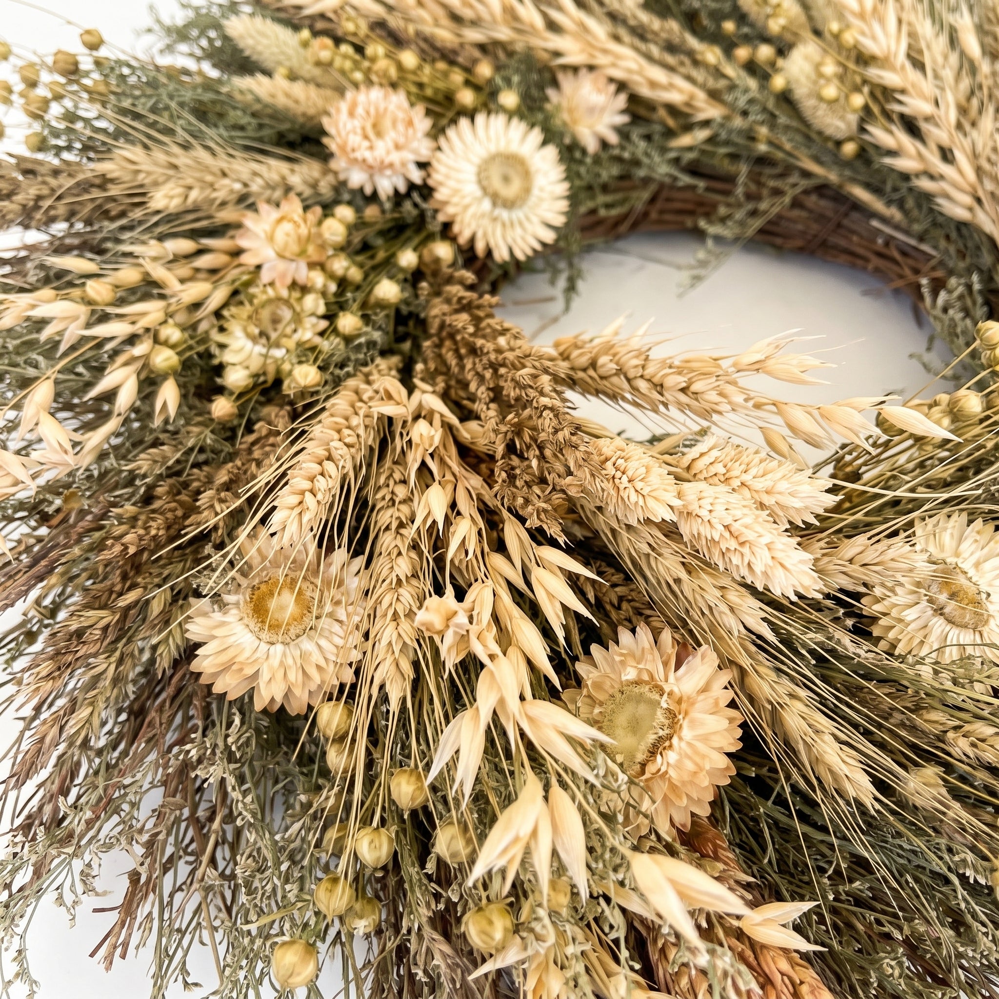 A dried flower and grass wreath made from natural wheat stalks, strawflowers, and small pale blooms on a light neutral background.