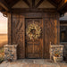 A rustic wooden front door with carved wheat motifs on the door panels, set in a stone-and-wood porch with a dried floral wreath hanging on the door.