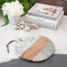 Round marble and wood cutting board on a table with decorative books and flowers.