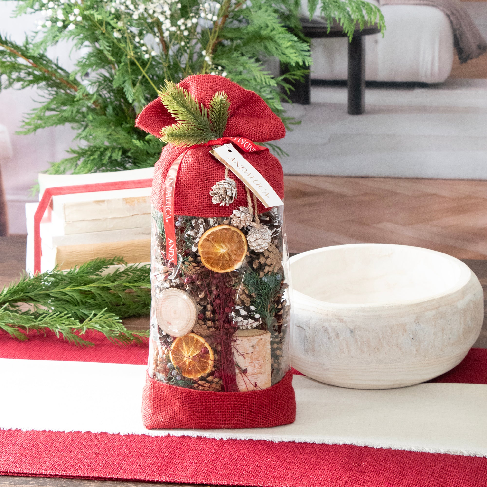 Holiday Memories Burlap Pinecone Bag is a decorative potpourri bag with red burlap fabric on the top & bottom. The bag is clear in the middle allowing it to act as a window to the content of pine cones, oranges, and other botanicals. Shot on a table top setting.