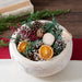 Close up of Holiday Memories Burlap Pinecone bag is displaed in a white wood bowl. Shot in a room setting with an open small journal with pen on it.