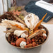 A decorative autumn arrangement in a shallow round bowl, featuring dried corn husks, wheat stalks, pinecones, acorns, small berries, and a small gourd, styled on a tabletop with books and a dark navy wallet in the background.