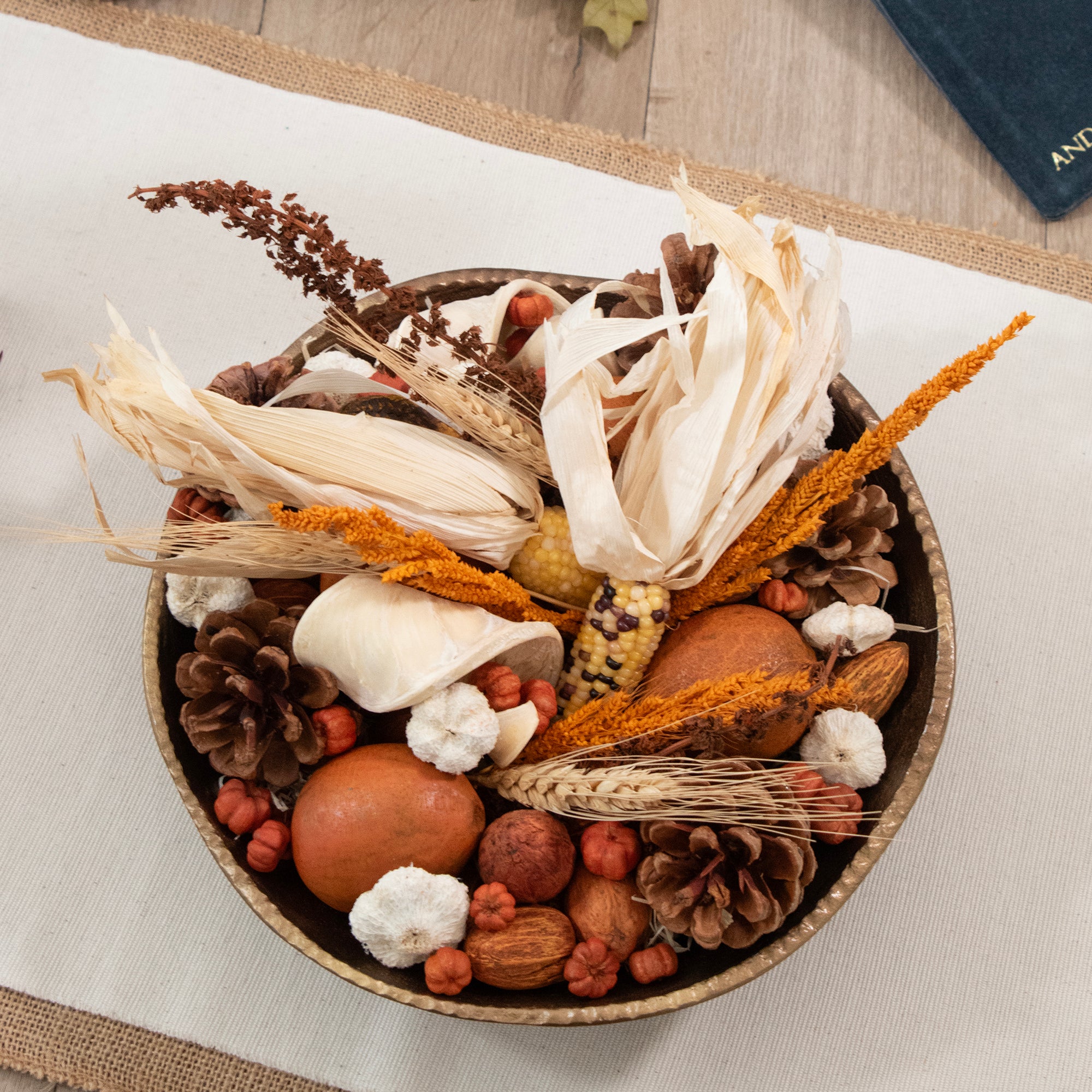 A decorative autumnal arrangement in a round woven bowl, featuring dried corn husks, orange grasses, pinecones, acorns, small red berries, and white flower buds on a light fabric surface.