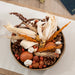 A decorative autumnal arrangement in a round woven bowl, featuring dried corn husks, orange grasses, pinecones, acorns, small red berries, and white flower buds on a light fabric surface.