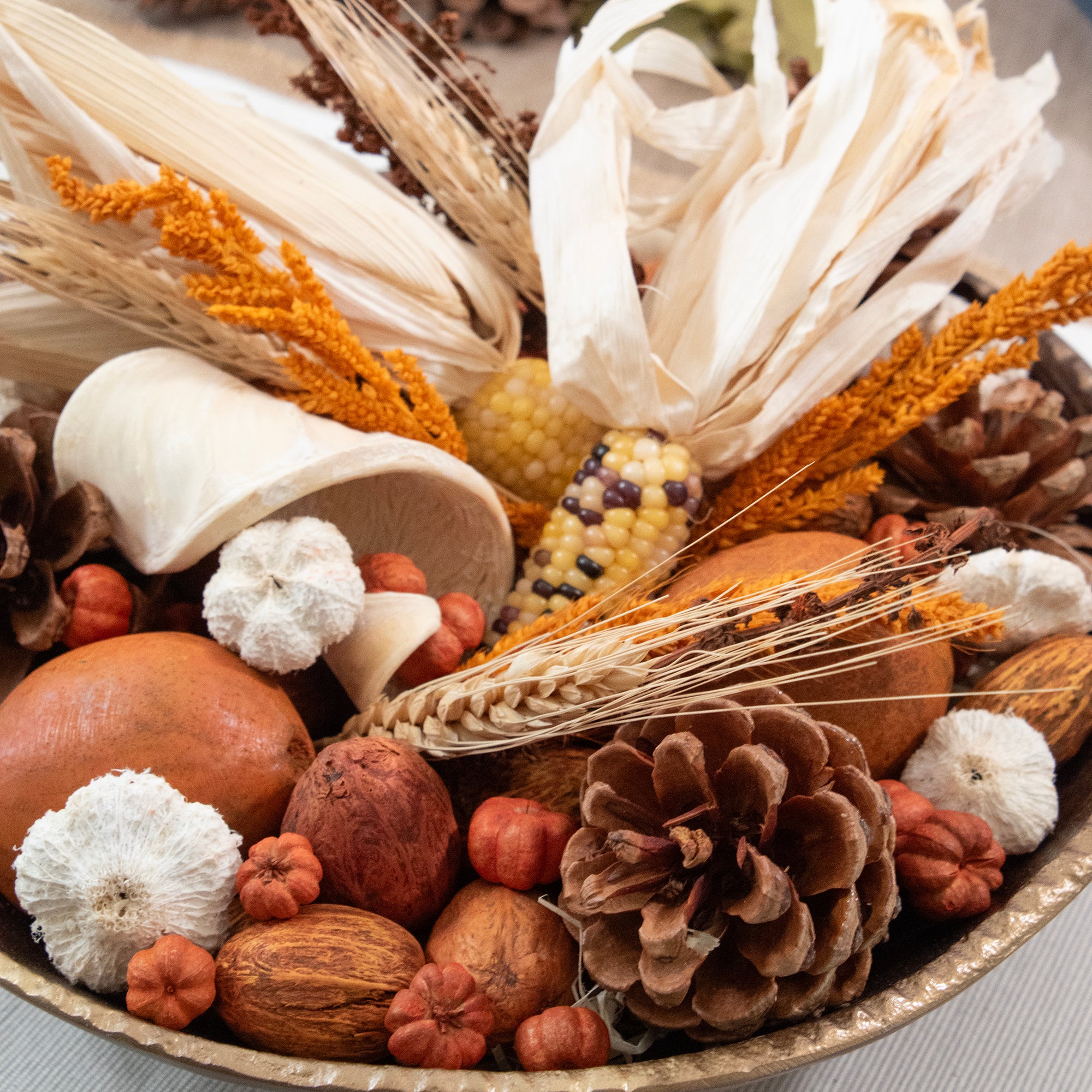 A close-up of a fall harvest arrangement in a bowl, featuring dried corn, corn husks, wheat stalks, pine cones, acorns, small pumpkins, and white mushrooms on a light surface.