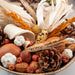 A close-up of a fall harvest arrangement in a bowl, featuring dried corn, corn husks, wheat stalks, pine cones, acorns, small pumpkins, and white mushrooms on a light surface.