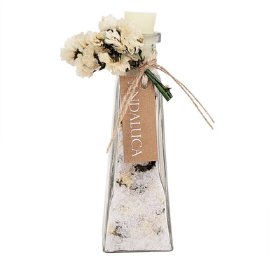 A clear glass bottle with a tapered shape, filled with white bath salts or sand, decorated with dried cream flowers, a brown kraft paper tag reading 'SALTUCA', and a small white candle at the top, shown on a white background.