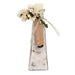 A clear glass bottle with a tapered shape, filled with white bath salts or sand, decorated with dried cream flowers, a brown kraft paper tag reading 'SALTUCA', and a small white candle at the top, shown on a white background.
