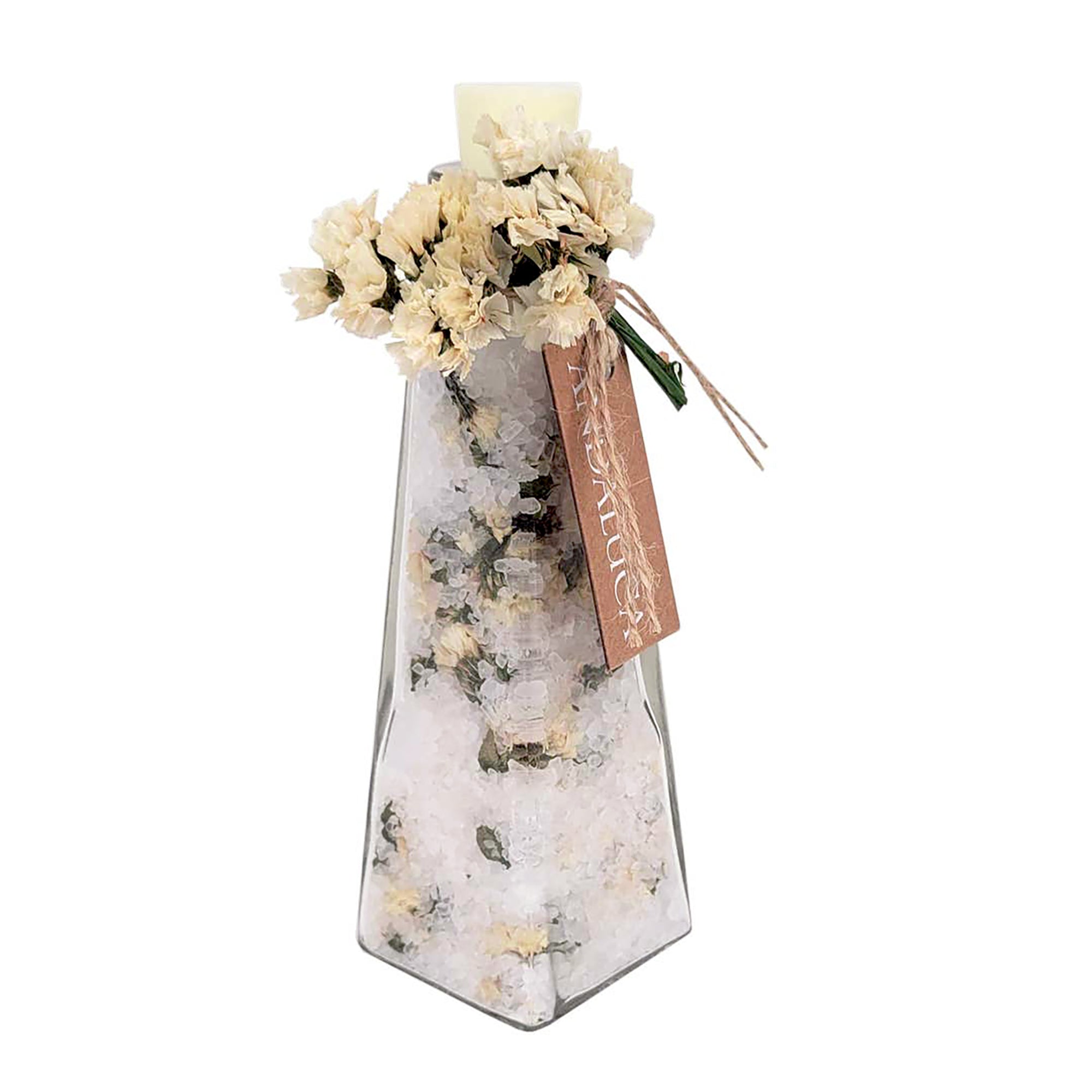 A tall, narrow clear glass vase filled with white bath salts and dried flowers, topped with a cream-colored candle and a brown paper tag reading 'MISTURE'.