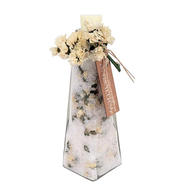 A tall, narrow clear glass vase filled with white bath salts and dried flowers, topped with a cream-colored candle and a brown paper tag reading 'MISTURE'.