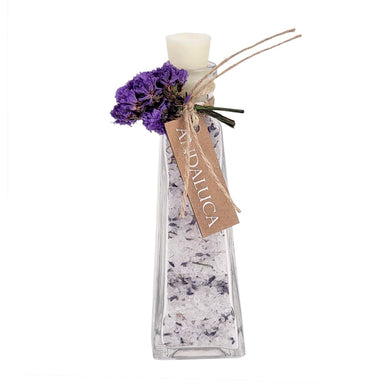 A clear glass bottle filled with white bath salts speckled with purple, topped with a white candle and decorated with dried purple flowers and a kraft paper tag reading 'ANDALUCA', on a white background.