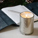 Mercury Candle Cup lit on a table top surface with an open book and pen in the background.