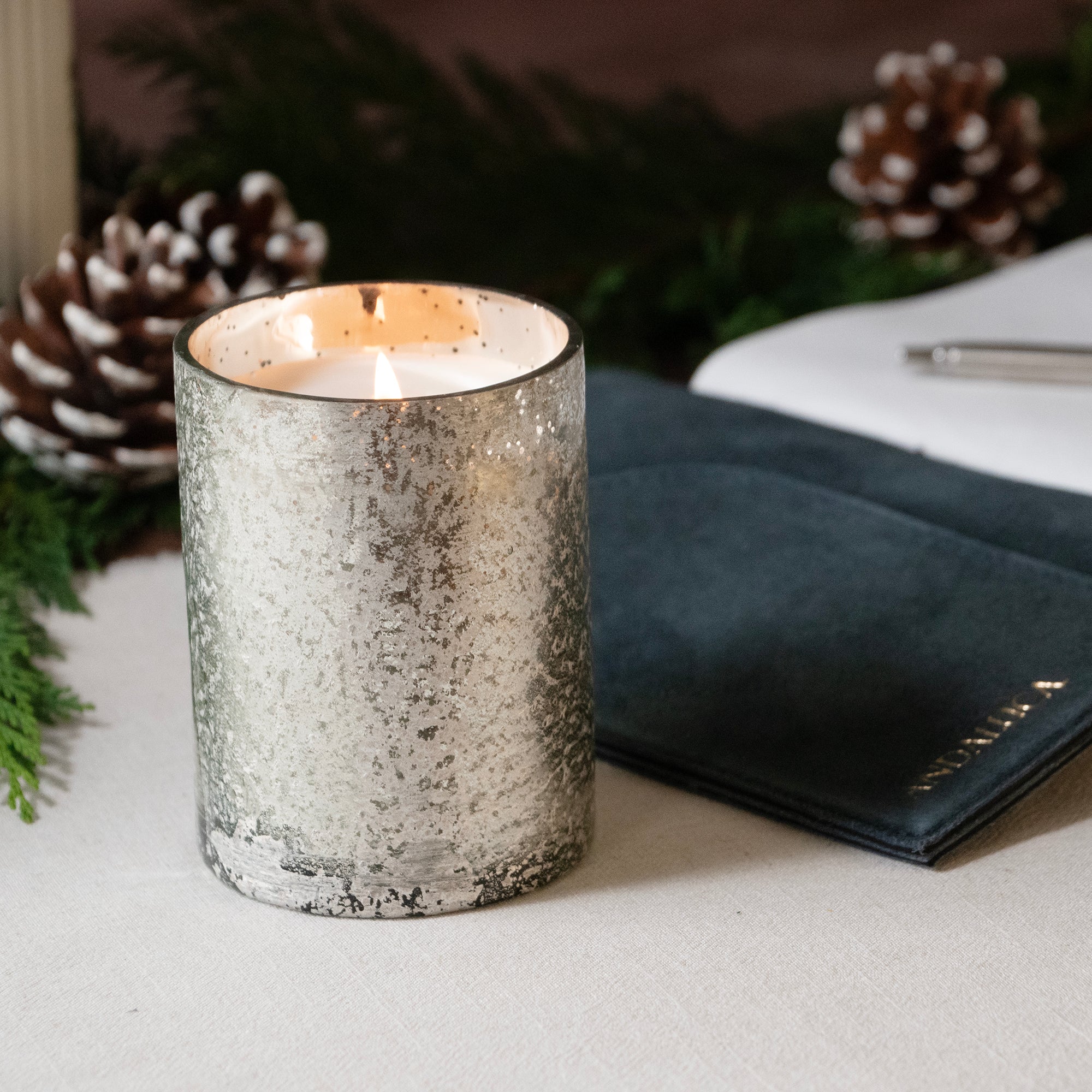 Mercury Candle Cup lit on a table top surface with an open book and pen in the background.