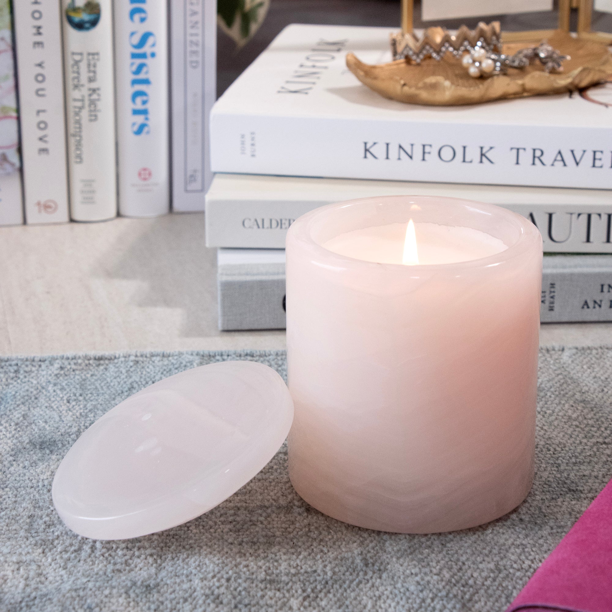 A lit pale pink pillar candle with its translucent lid resting beside it on a gray textured surface, with stacked books in the background.