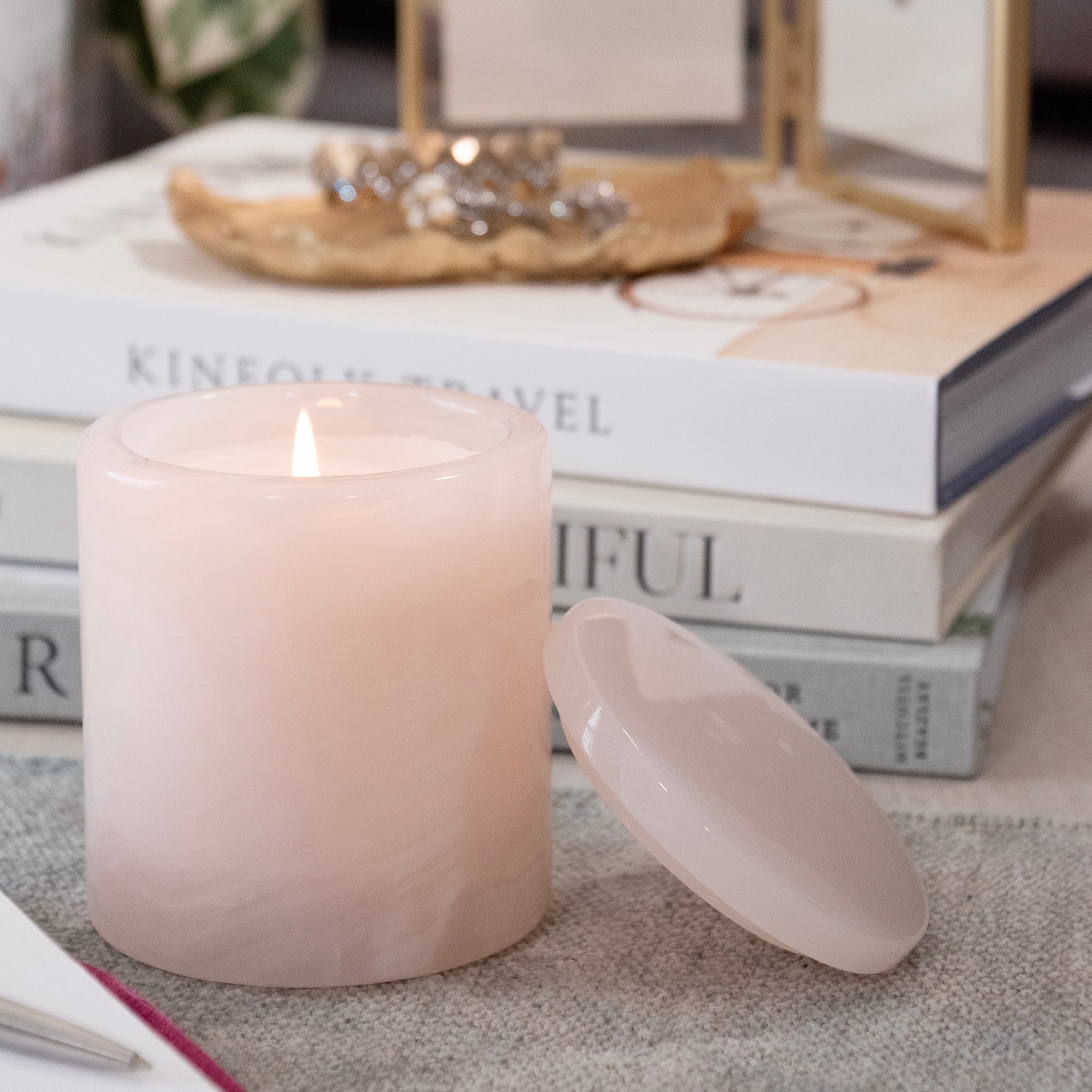 A pale blush pink marbled candle in a matching glass jar with its lid leaning beside it on a textured surface, with stacked books and a decorative tray in the softly blurred background.