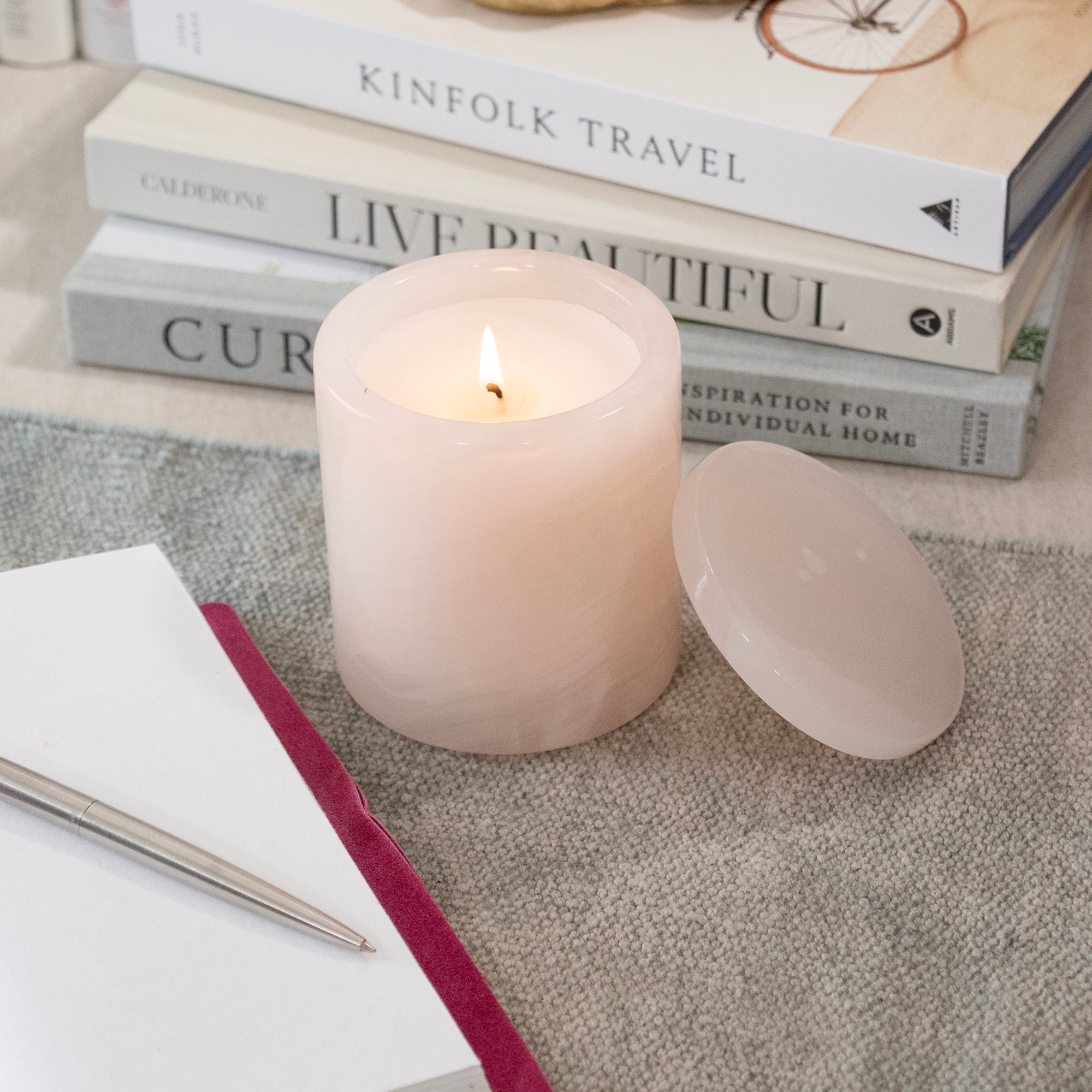 A pale blush pink marbled candle lit sits on a textured surface, with stacked books and a decorative tray in the softly blurred background.