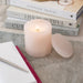 A pale blush pink marbled candle lit sits on a textured surface, with stacked books and a decorative tray in the softly blurred background.
