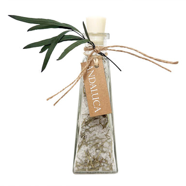 A clear glass bottle with a white stopper, filled with coarse white salt crystals and dried botanicals, tied with twine and a kraft paper tag reading ANDALUCA, with green leaves attached, on a white background.