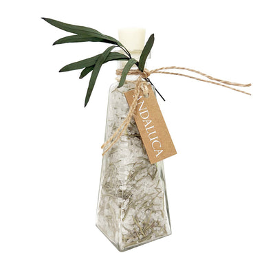 A clear glass bottle filled with dried white botanicals and tied with twine, featuring a kraft paper tag that reads 'ANDALUCA' and green leaves at the top, shown on a white background.
