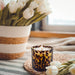 A lit candle in a dark amber-brown tortoise print glass holder sits on a woven placemat on a striped blanket, with white tulips in a striped basket and more tulips blurred in the background near a window.