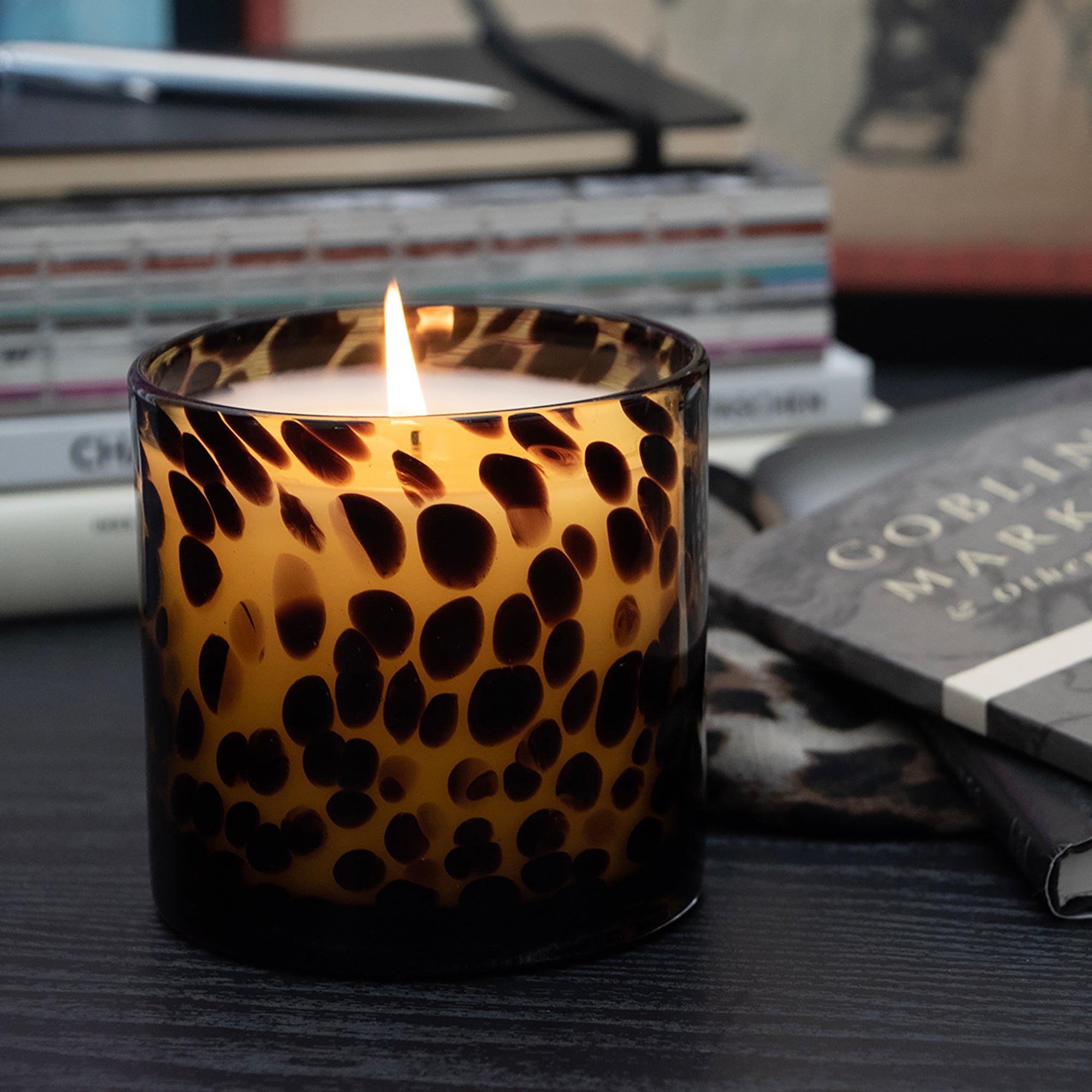 A  lit Tortoise Glass candle, set on a dark tabletop with books in the background.