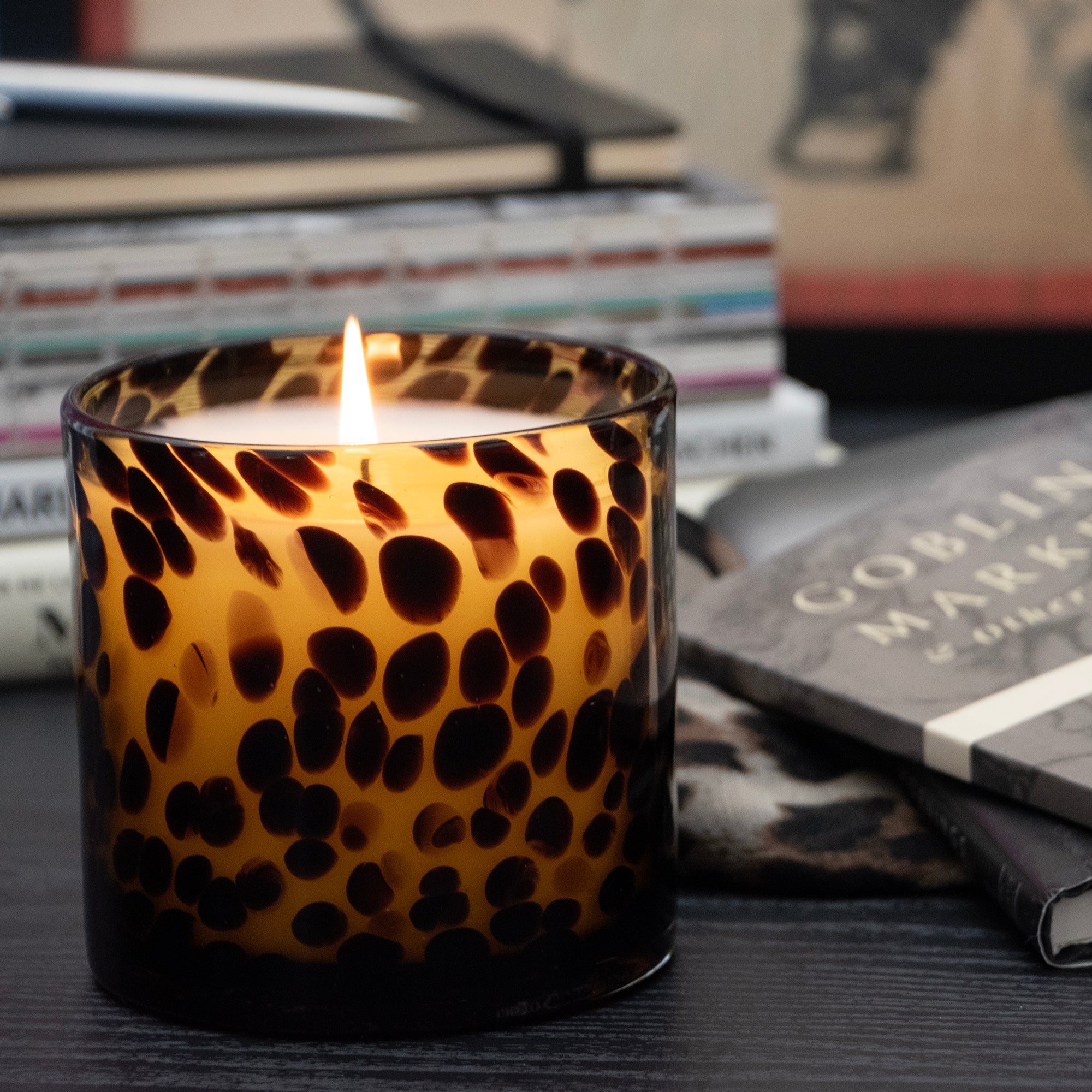 A lit Tortoise Glass candle, set on a dark tabletop next to books.