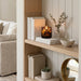 A lit Tortoise Glass candle sits on a wooden shelf beside stacked books, a small vase with eucalyptus, and a ceramic bowl in a styled interior setting.