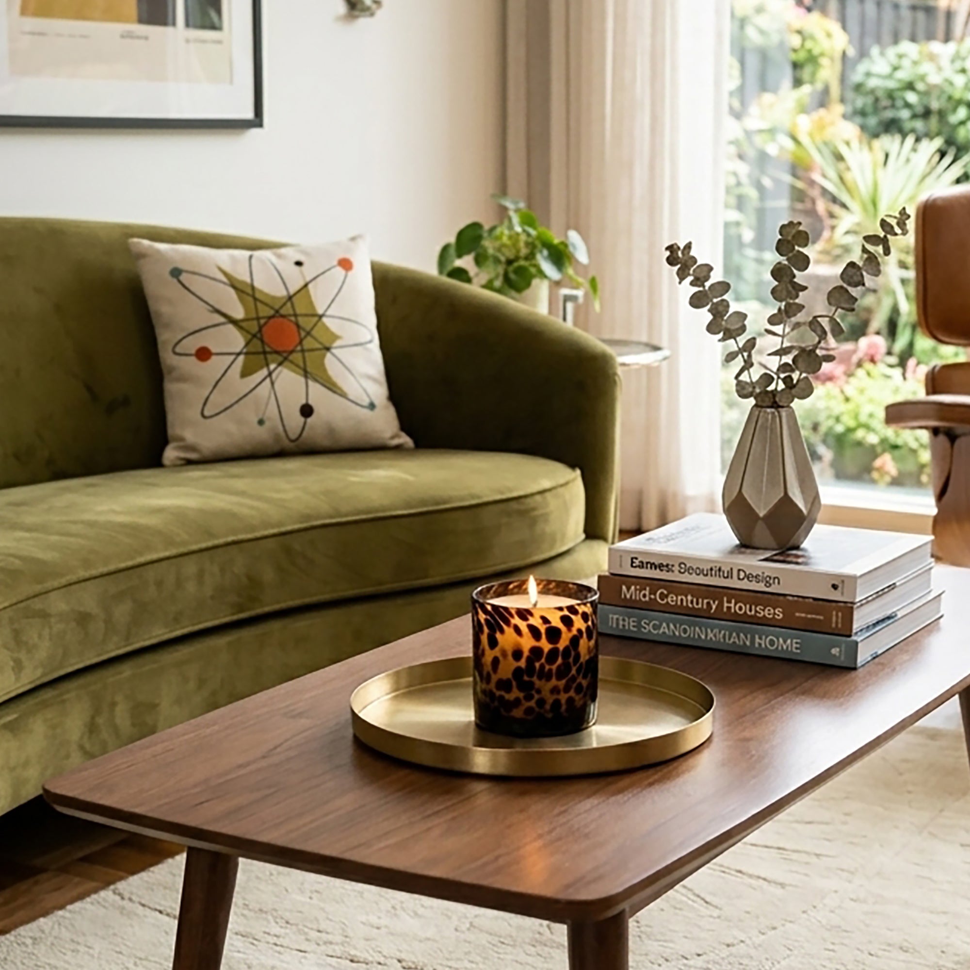 A lit Tortoise Glass candle sits on a round brass tray on a wooden coffee table in a styled living room with an olive green sofa and a window view.